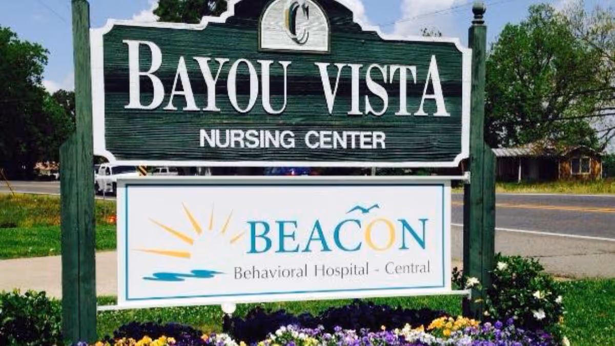 Wooden roadside sign reading "Bayou Vista Nursing Center" with a lower panel for "BEACON Behavioral Hospital - Central" set by a road and flowerbed.
