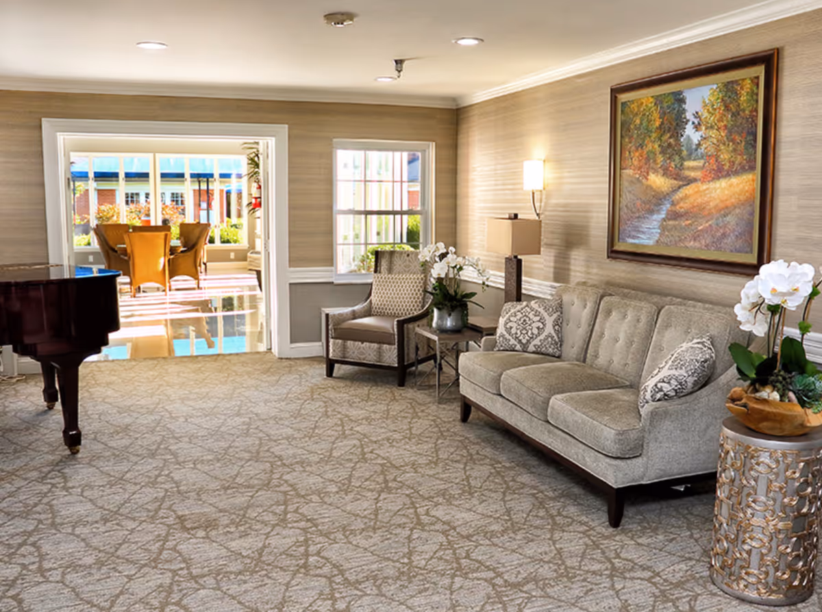 A cozy living room area with a gray tufted sofa adorned with patterned pillows, a matching armchair, a side table with a lamp and white orchids, and a decorative round table with a white orchid plant. A framed painting of a scenic autumn landscape hangs on the wall. In the background, a sunlit dining area with a table and chairs is visible through an open doorway. A grand piano is partially visible on the left side of the room.