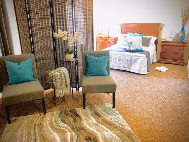 A cozy bedroom area with a bed featuring white and blue bedding, flanked by two wooden nightstands with decorative items. In the foreground, there are two gray chairs with teal pillows, a small round table with a plant, and a patterned rug on the carpeted floor. A folding screen is positioned behind the chairs.