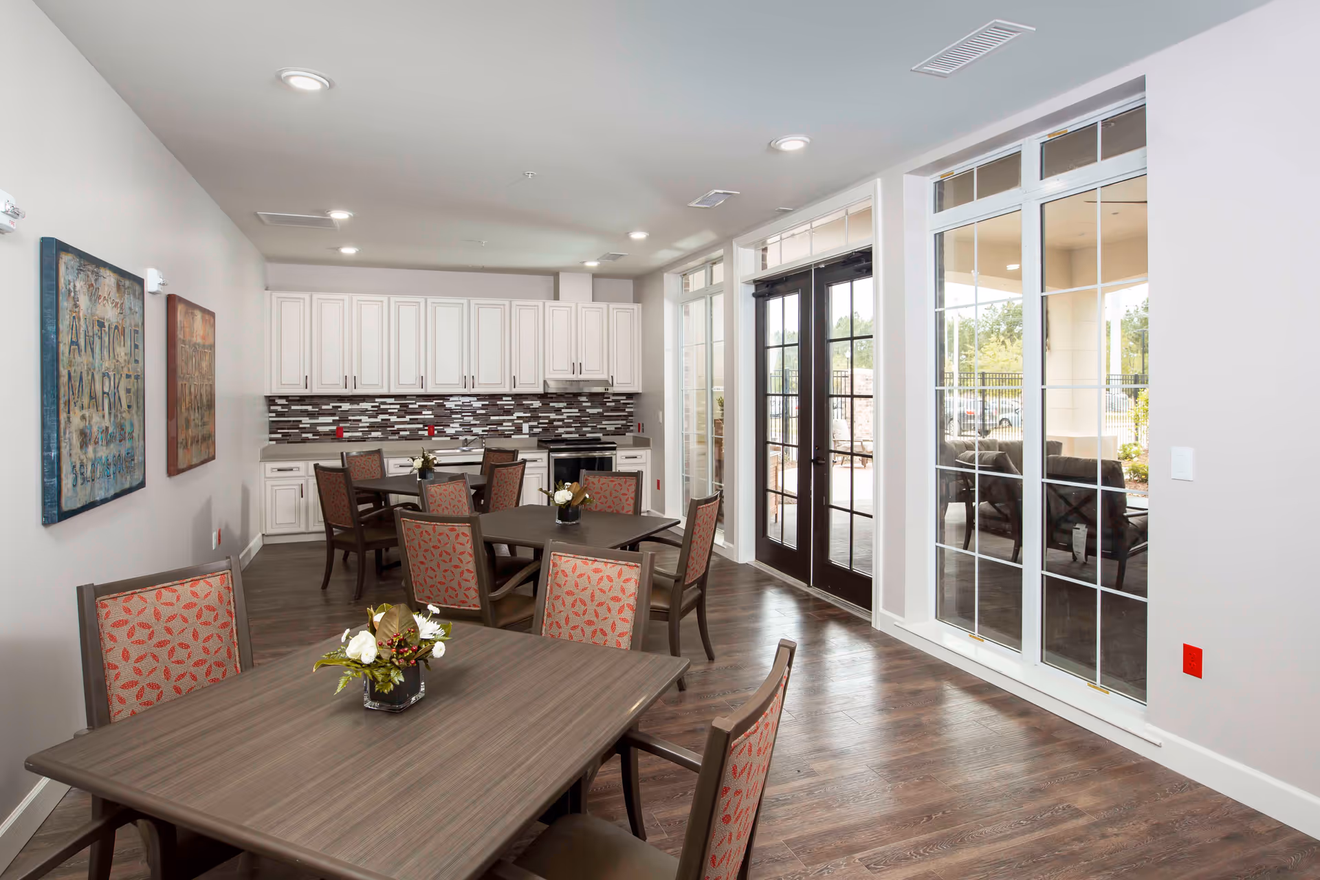 Bright communal dining area with wooden tables and patterned chairs, a kitchenette along the back wall, and large glass doors leading outside.