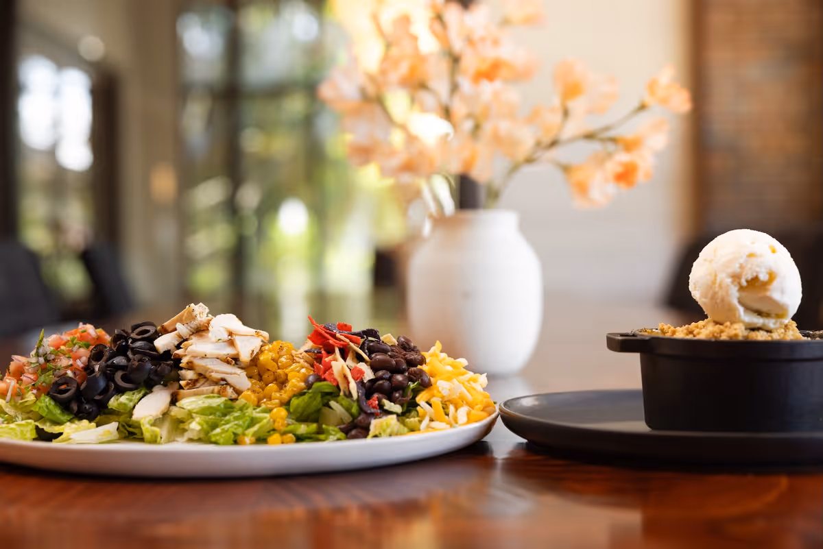 A plated salad and a dessert with ice cream on a wooden table in a softly lit dining area with a vase of flowers in the background.