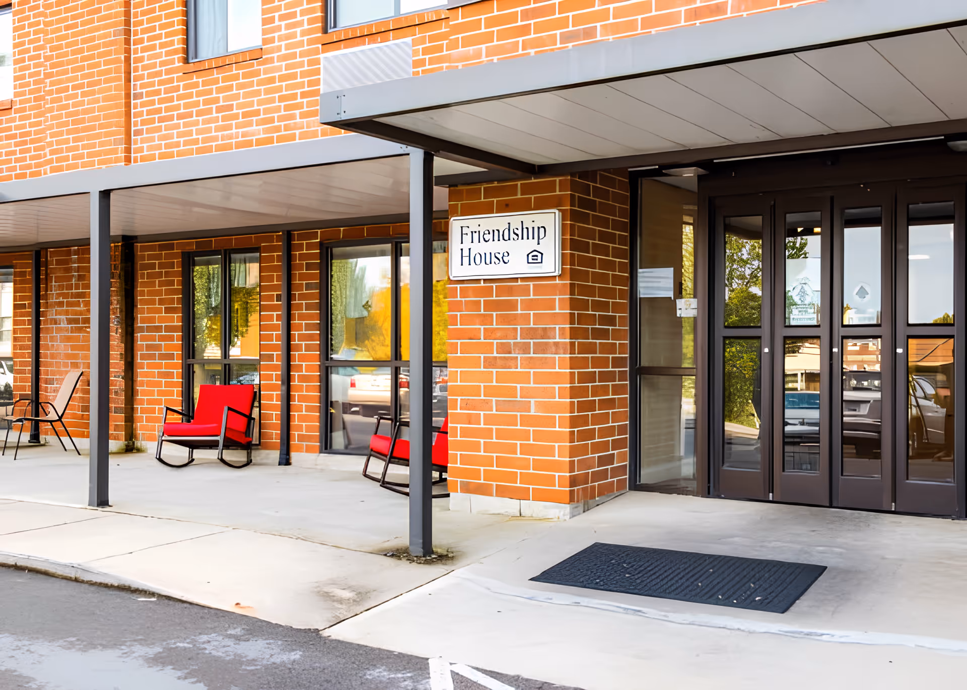 Brick-front entrance with a 'Friendship House' sign, glass doors, and red chairs on a covered porch.