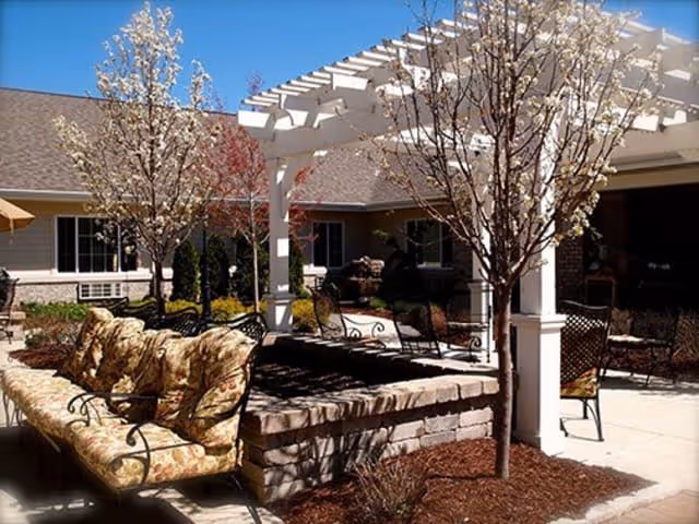 Outdoor seating area with cushioned benches and chairs under a white pergola surrounded by small trees and landscaping in a senior living facility courtyard.