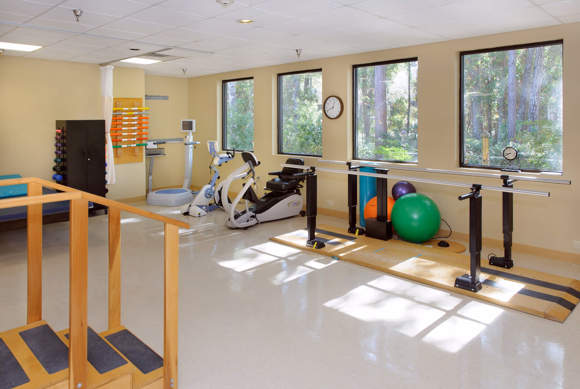 A bright physical therapy room with large windows showing a wooded outdoor view. The room contains exercise equipment including stationary bikes, parallel bars for walking practice, colorful exercise balls, a set of stairs with handrails, and a rack of small dumbbells. The walls are light yellow and the floor is a light-colored linoleum.