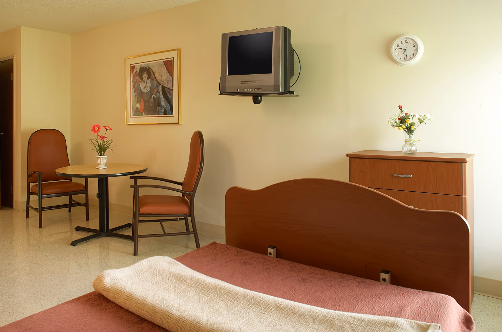 A senior living facility bedroom with a wooden bed covered with a pink blanket and a beige throw. There is a wooden dresser with a vase of flowers on top, a wall-mounted TV, a round table with two chairs, a framed painting on the wall, and a clock showing the time as 3:10.