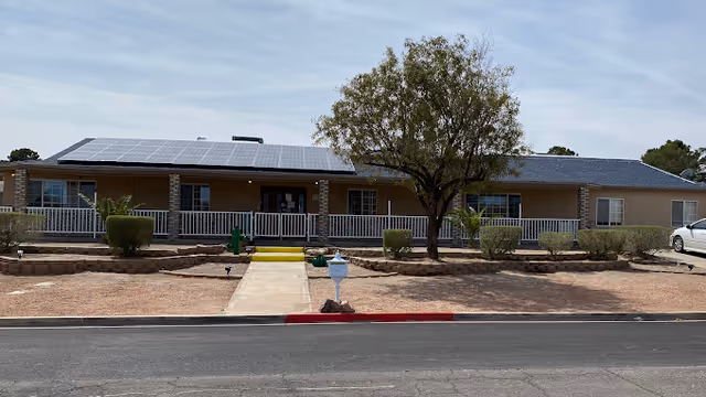 Single-story building with a covered porch and white railing, solar panels on the roof, a tree and trimmed bushes in front, a concrete walkway leading to the entrance, and a mailbox near the street.