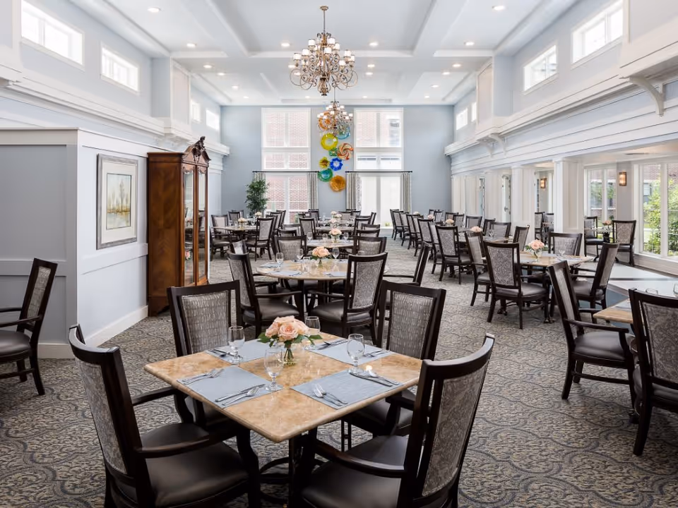 A spacious and elegant dining room with multiple tables set for four, each adorned with a small vase of pink roses, placemats, glassware, and silverware. The room features high ceilings with chandeliers, large windows allowing natural light, light blue walls, and decorative wall art. The carpet has a patterned design, and there is a wooden cabinet along one wall.