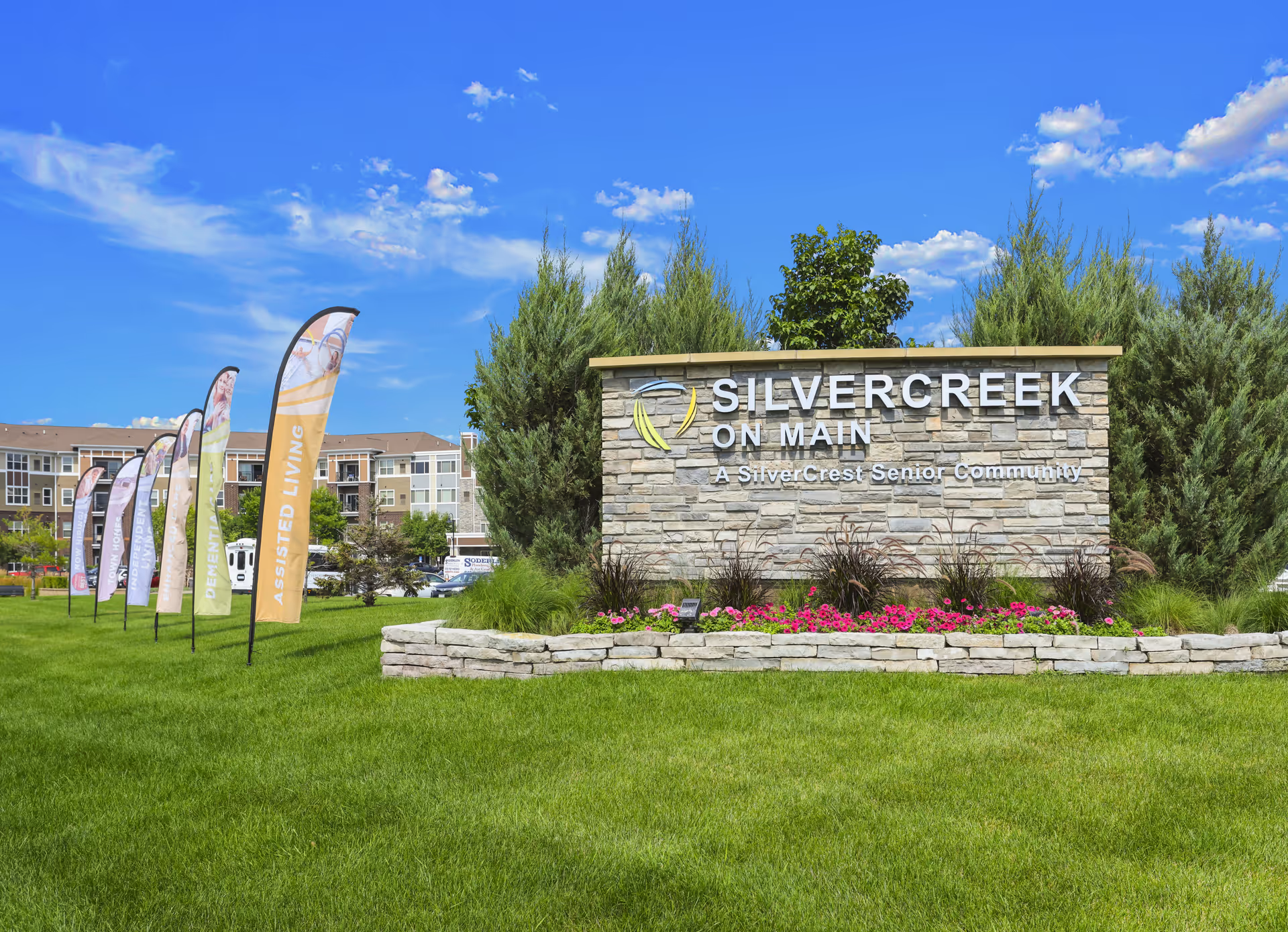Outdoor view of SilverCreek on Main senior living community sign made of stone with landscaping and flowers in front, multiple colorful flags advertising services like assisted living, and a large building in the background under a blue sky with scattered clouds.