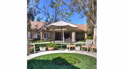 Outdoor courtyard area with a circular patch of green grass surrounded by a paved walkway. There are wooden chairs and benches arranged around the walkway, along with a large white patio umbrella providing shade. Trees and potted plants are visible, and a building with a tiled roof is in the background under a clear blue sky.