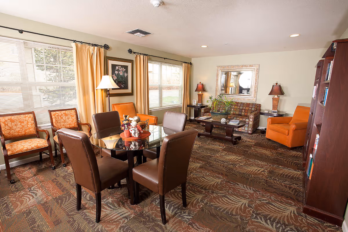 Sunlit common room with a round glass table and chairs, sofas and armchairs, lamps and bookshelves.