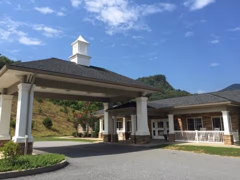 Exterior view of a senior living facility named The Hermitage showing a covered entrance with white columns and a cupola on the roof. The building has a brick and siding facade with a porch area and white railings. The background includes green hills and a partly cloudy blue sky.