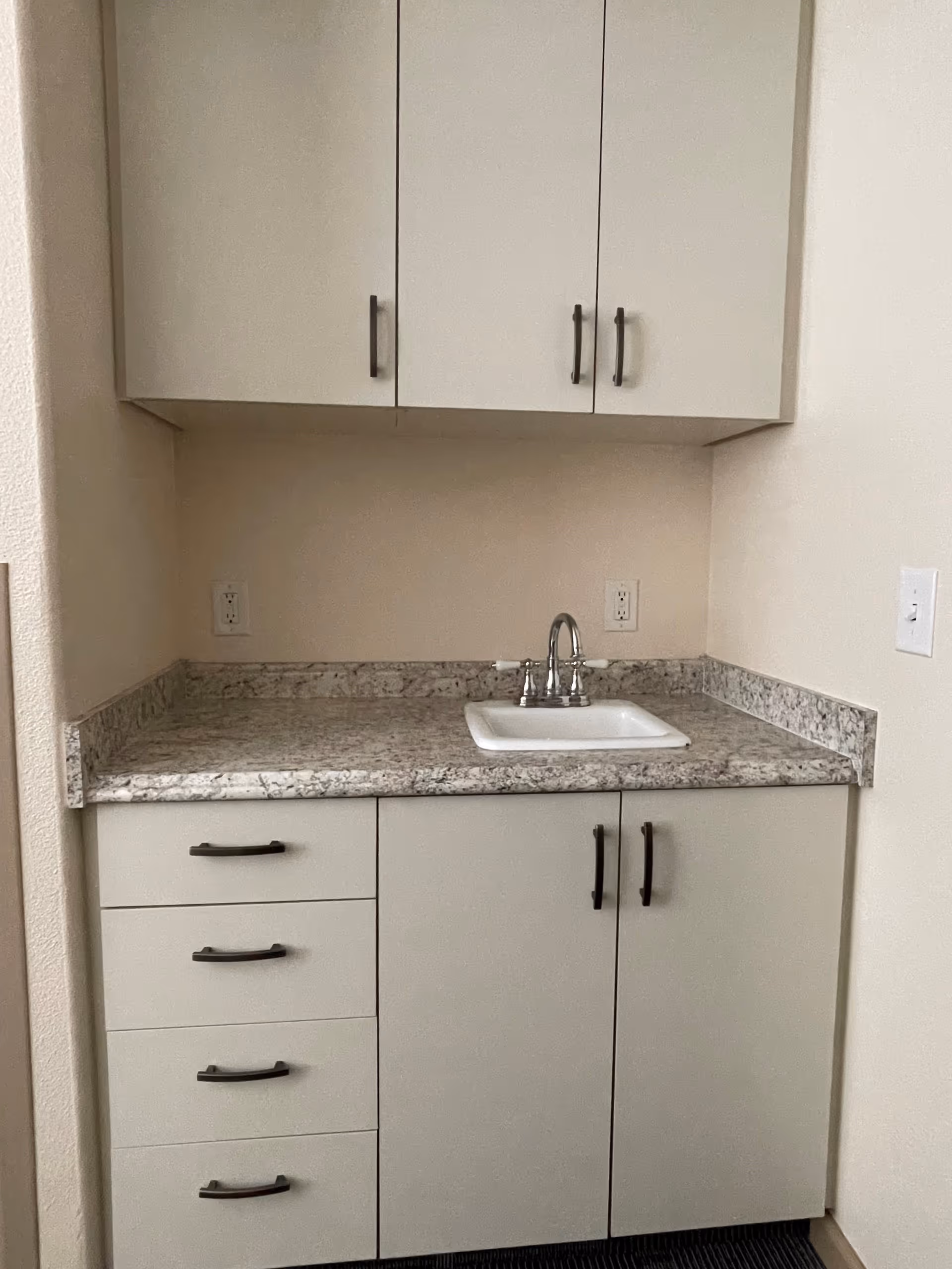 A small kitchenette area with a white sink and silver faucet set into a speckled granite countertop. There are white cabinets above and below the countertop with black handles. The walls are light-colored and there are electrical outlets on the backsplash.