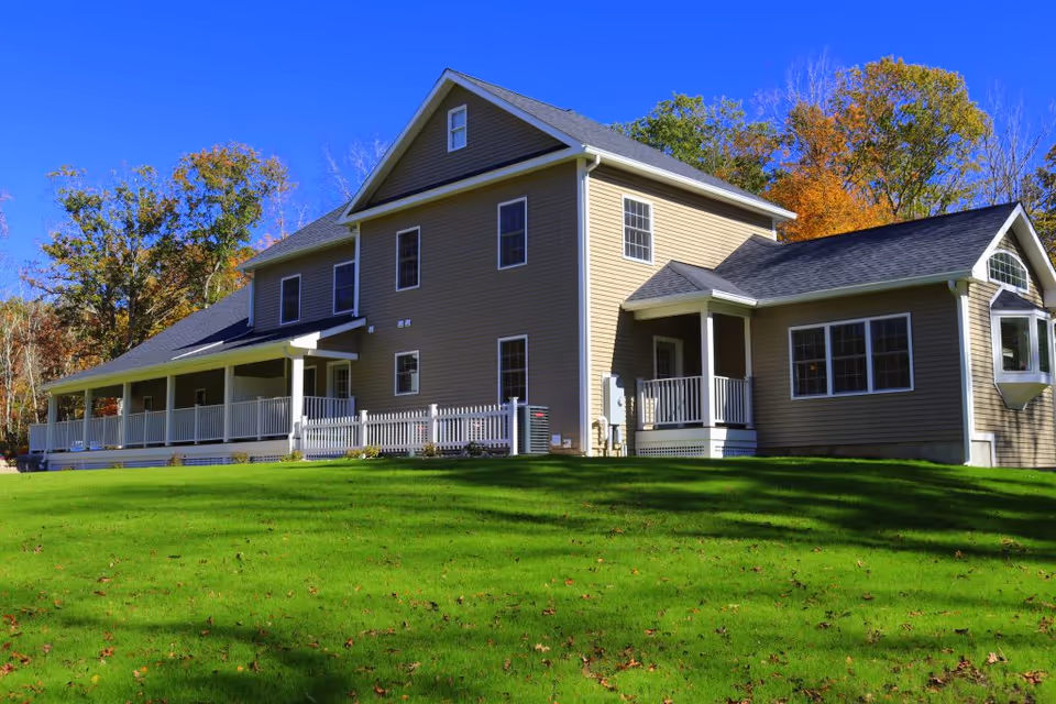 A large two-story residential care home with beige siding and a dark gray roof, surrounded by green grass and trees with autumn foliage under a clear blue sky.