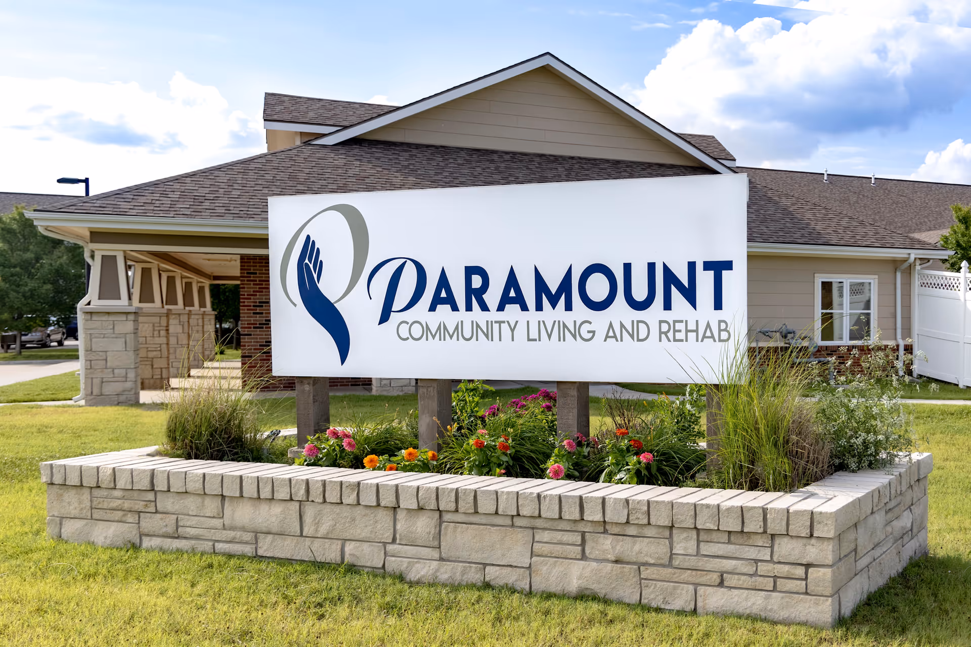 Outdoor view of a sign for Paramount Community Living and Rehab in front of a building with a stone planter containing flowers and greenery.