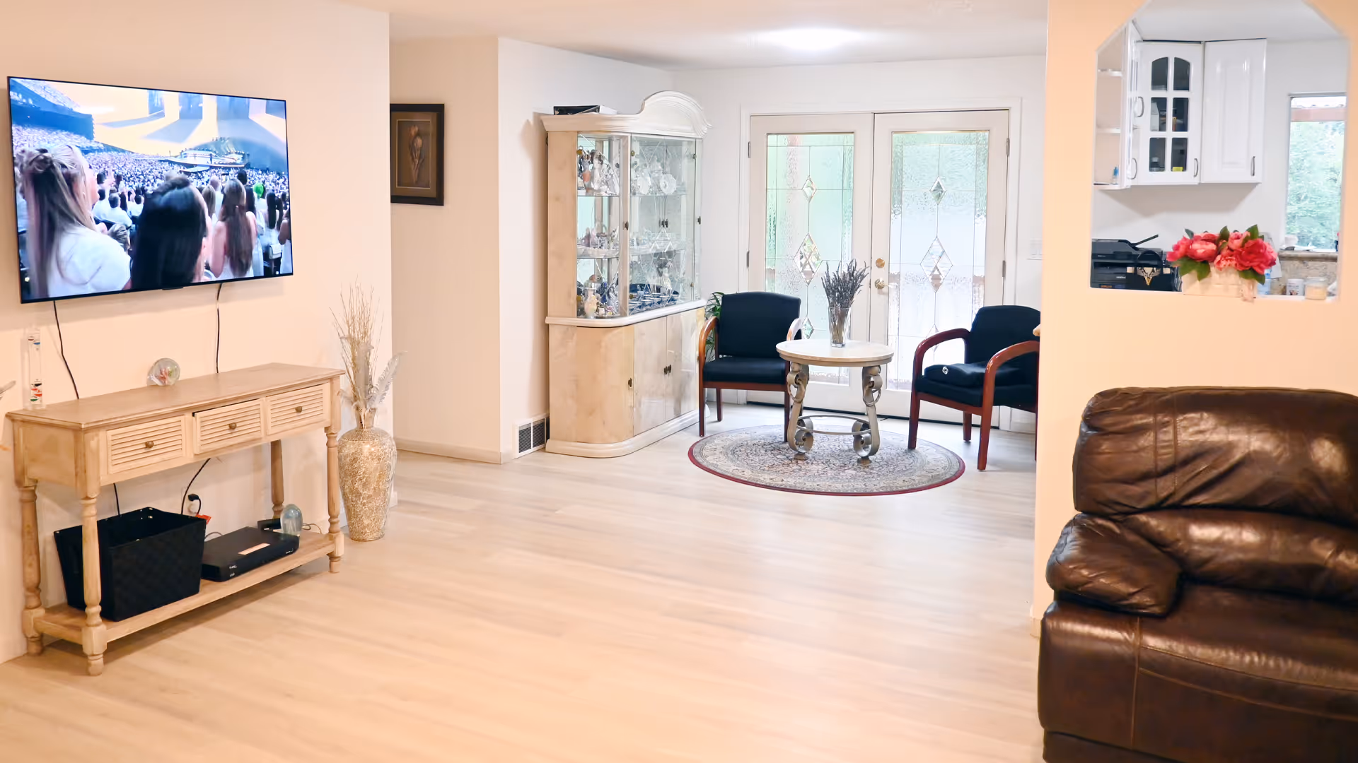Bright living room in a senior home with a wall-mounted TV, a wooden console, glass display cabinet, a small seating area around a round table, and a brown leather recliner in the foreground.