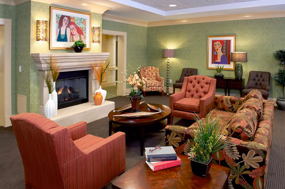 A cozy living room with a fireplace, featuring a floral patterned sofa, two red armchairs, and two brown tufted chairs. The room has green wallpaper, framed artwork on the walls, a round wooden coffee table with decorative items, and a side table with books and a plant. Soft lighting comes from wall sconces and lamps.