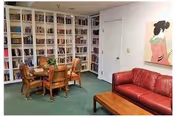A cozy room with a red leather sofa, a wooden coffee table, a round wooden table with four chairs, and a large white bookshelf filled with books along one wall. There is a door and a colorful painting on the wall.