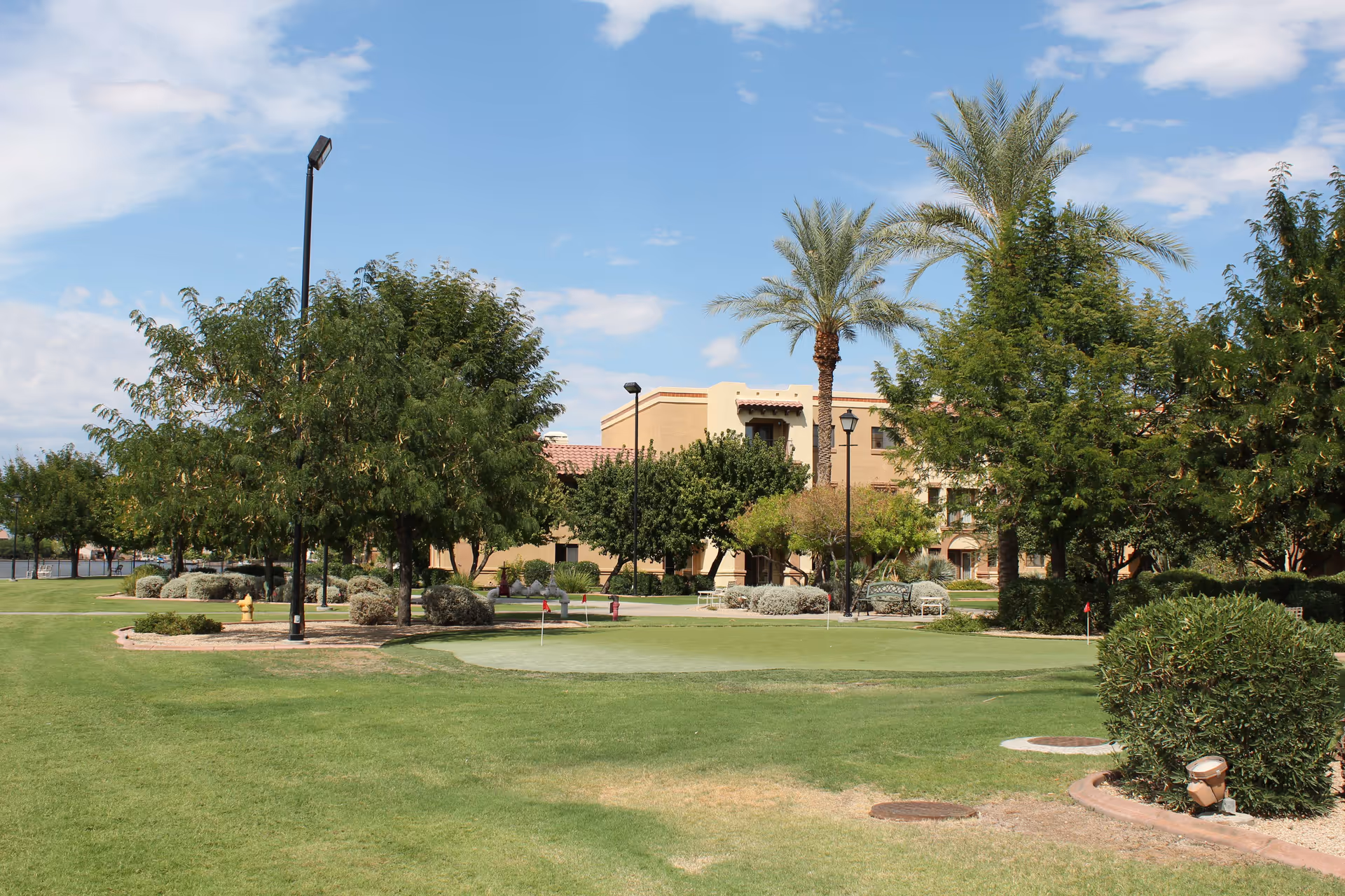 Outdoor view of a senior living facility with a well-maintained green lawn, palm trees, other trees, bushes, and a putting green. The building with beige walls and red-tiled roof is visible in the background under a partly cloudy blue sky.