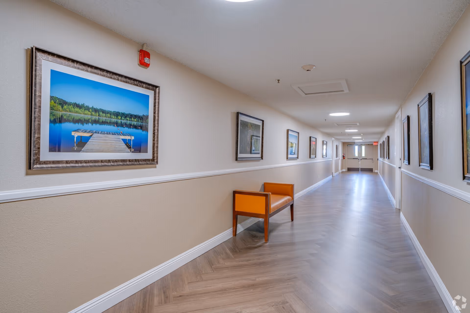 A long, well-lit hallway with light-colored walls and wood-patterned flooring. The walls are decorated with framed landscape photographs, and there is an orange bench along one side. The hallway leads to a set of double doors at the far end.