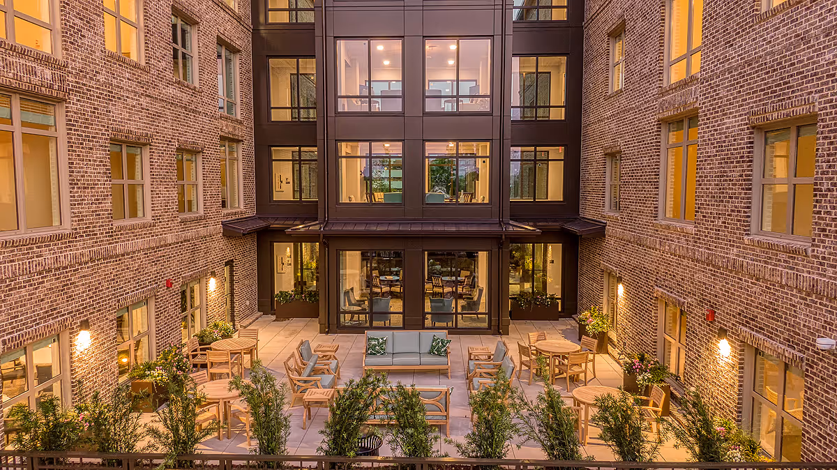 Outdoor courtyard area of a senior living facility with multiple round wooden tables and chairs arranged on a tiled patio. There is a central seating area with a sofa and armchairs with cushions. The courtyard is surrounded by a brick building with multiple windows, some of which are lit from inside. There are plants and greenery along the edges of the patio, and warm lighting illuminates the space.
