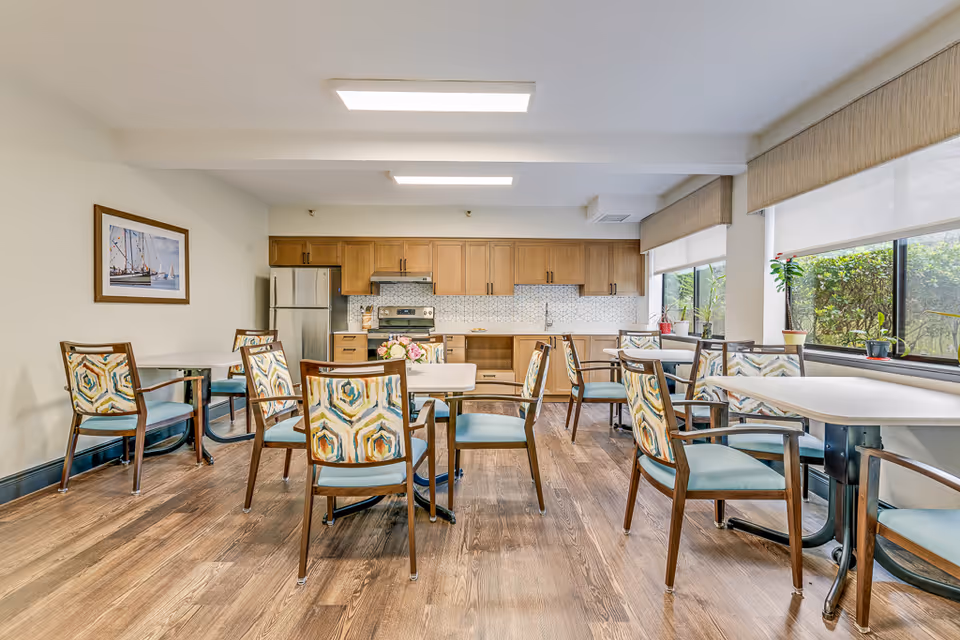 A bright dining room with several tables and chairs featuring colorful patterned backs and light blue seats. The room has large windows with beige valances letting in natural light, wooden flooring, and a kitchen area with wooden cabinets, a stainless steel refrigerator, stove, and a tiled backsplash. There are potted plants on the windowsill and a framed picture of a sailboat on the wall.