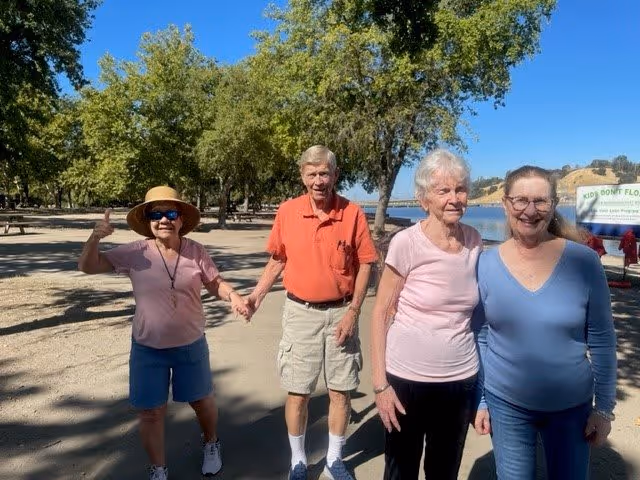 Four elderly people standing outdoors on a sunny day near a body of water with trees in the background. One woman on the left is wearing a hat and sunglasses, giving a thumbs-up. The group appears happy and relaxed.