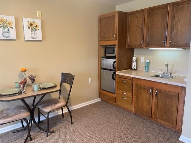 Small kitchenette area with wooden cabinets, a sink, a microwave, and a mini refrigerator. Adjacent to the kitchenette is a small dining table set for two with plates, mugs, and a small flower vase. The walls are beige with two sunflower paintings hanging above the table.