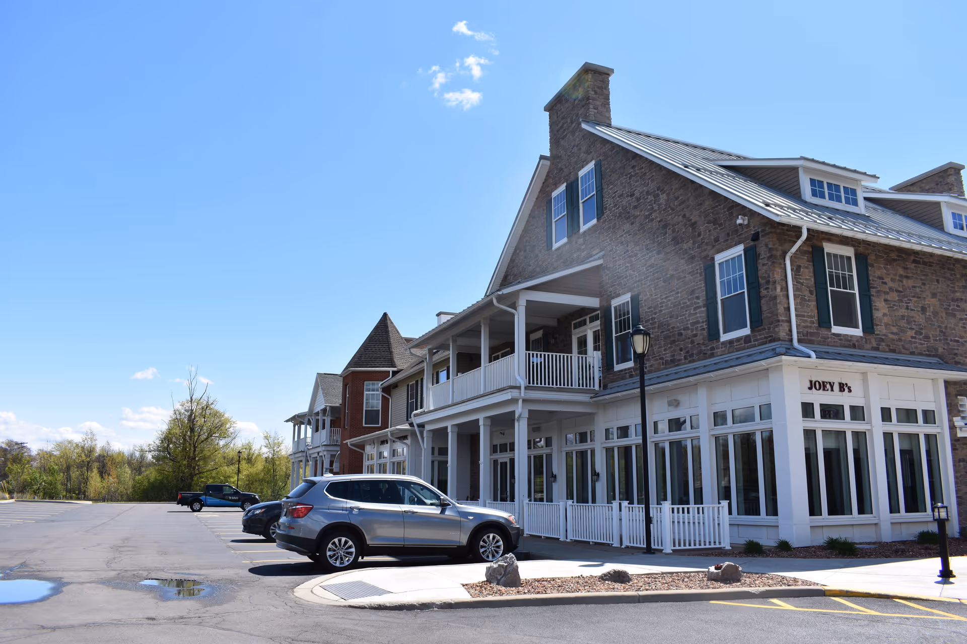 Exterior front of a multi-story brick senior living building with a covered porch, storefront sign reading "JOEY B's", parked cars, and a clear blue sky.
