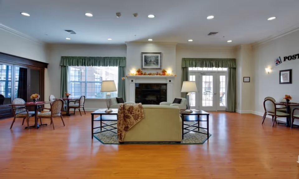 A spacious and well-lit living room area with wooden flooring, a central beige sofa facing a fireplace, two side tables with lamps, and several small round tables with chairs near large windows and glass doors. The room has green curtains and a framed picture above the fireplace.