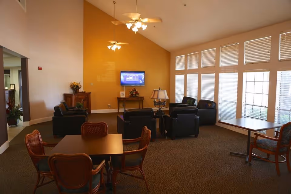A spacious living room area with a high ceiling and large windows with blinds allowing natural light. The room features a yellow accent wall with a mounted flat-screen TV, several black leather armchairs arranged around a small table with a lamp, and two wooden tables with chairs in the foreground. There are ceiling fans with lights hanging from the ceiling.