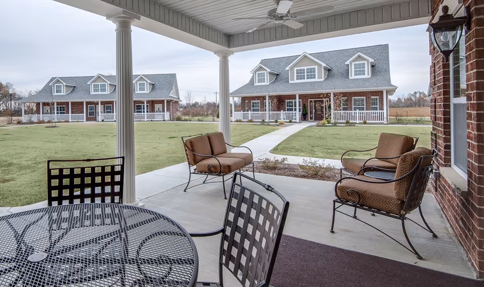 View from a covered patio with metal table and chairs and cushioned armchairs, looking out onto a green lawn with two large brick residential buildings with white trim and porches in the background under a cloudy sky.