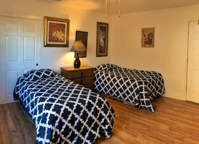 A bedroom with two twin beds covered in matching black and white patterned bedspreads. Between the beds is a wooden nightstand with a black lamp. The walls are decorated with framed artwork, and the floor is wooden.