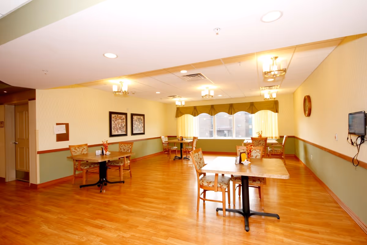 A bright dining room with wooden floors and several tables and chairs arranged neatly. The walls are painted in a two-tone style with a light yellow upper section and green lower section, separated by a wooden trim. There are framed artworks on one wall and a large window with green valances letting in natural light. Ceiling lights provide additional illumination.
