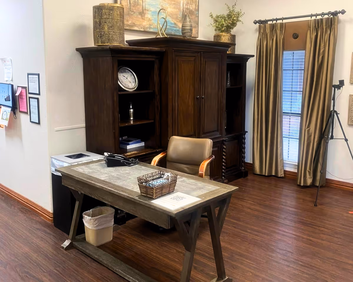 An interior office scene with a wooden desk and basket, a leather chair, a dark wood armoire, and curtains over a window.