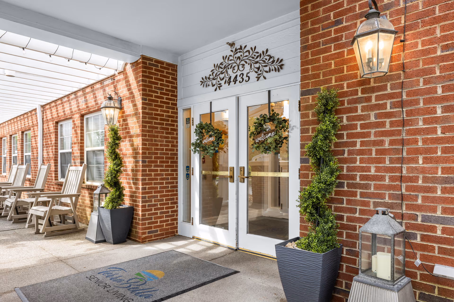 Front entrance of a brick senior living building featuring double glass doors with welcome wreaths, potted spiral topiaries, lanterns, a welcome mat, and a covered walkway with rocking chairs.