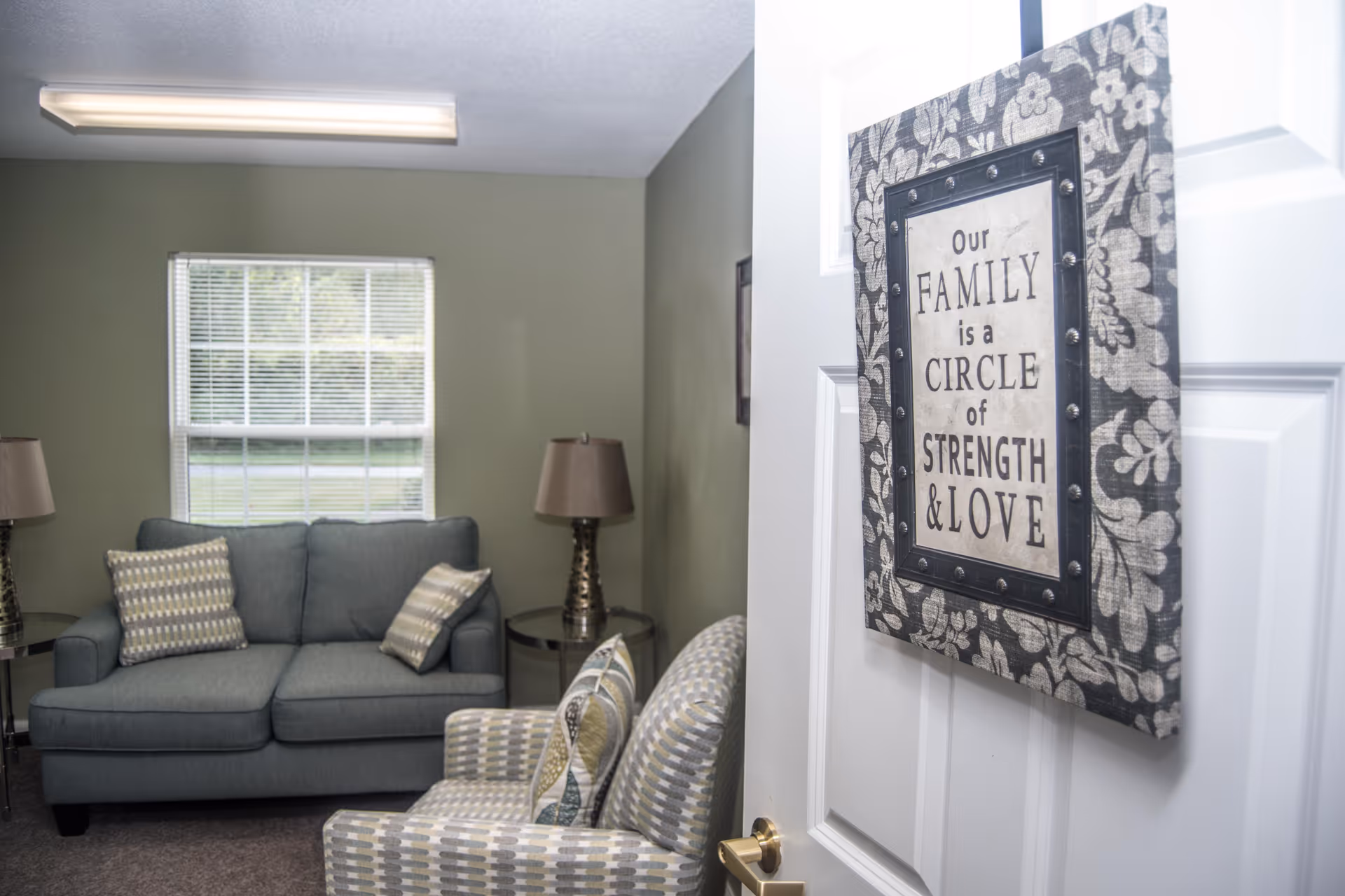 A cozy living room with a gray loveseat and two patterned armchairs, each with matching pillows. Two side tables with lamps flank the loveseat. A window with blinds lets in natural light. On the door in the foreground, there is a decorative sign that reads 'Our FAMILY is a CIRCLE of STRENGTH & LOVE.'
