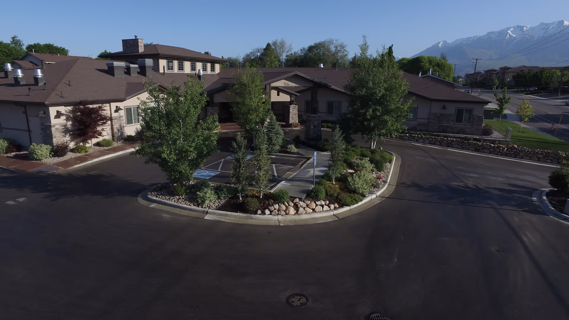 Front exterior view of Aspen Ridge of Utah Valley facility with a circular driveway, landscaped island with trees and shrubs, and a backdrop of mountains under a clear sky.