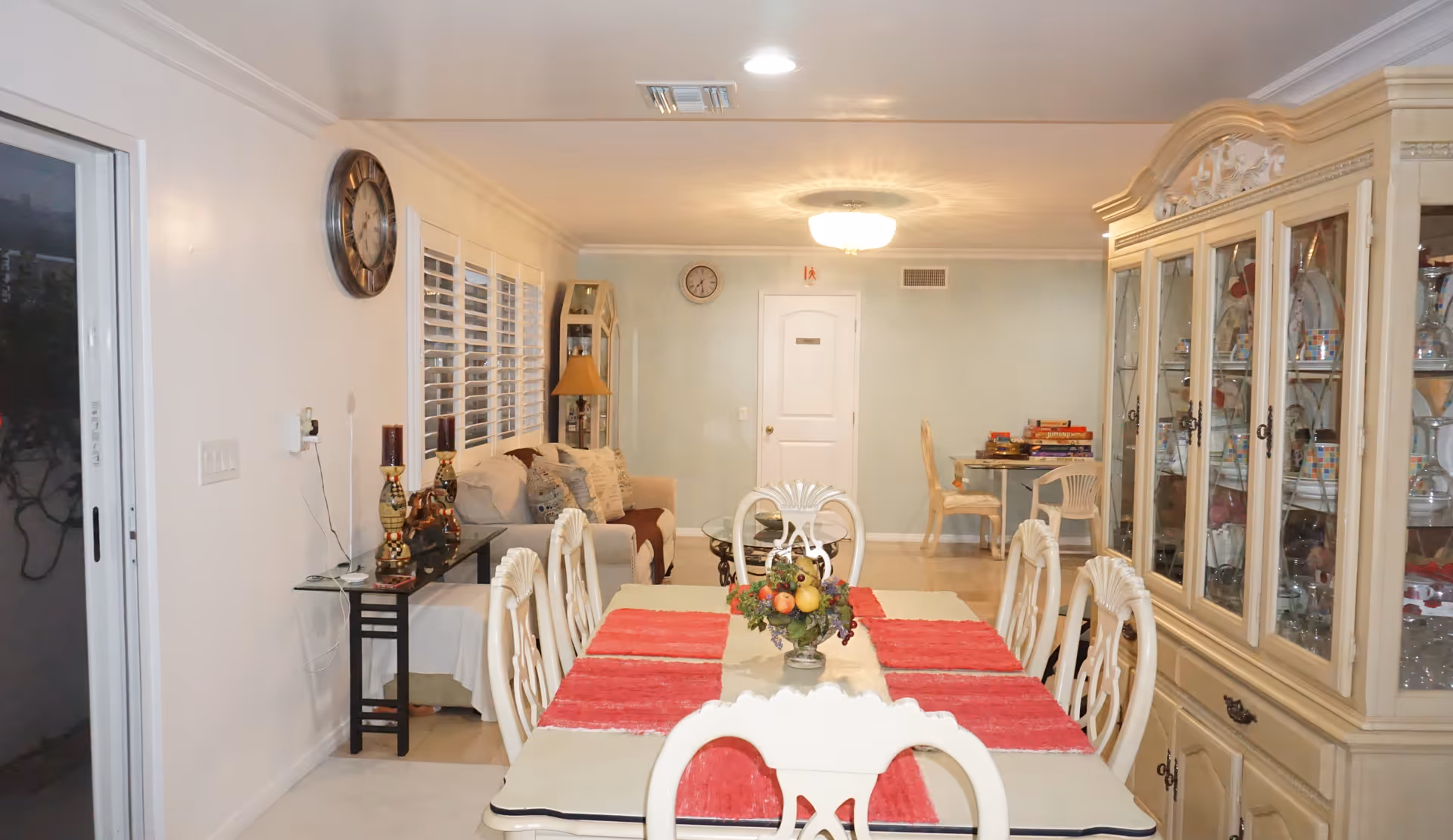 Interior view of a senior living facility showing a dining area with a table set with red placemats and a fruit centerpiece. Surrounding the table are white chairs with decorative backs. To the right is a large glass-fronted cabinet displaying dishes and glassware. In the background, there is a sitting area with a beige couch, a side table with lamps and decorative items, a clock on the wall, and a small table with chairs and board games. The walls are light-colored, and the room is well-lit with ceiling lights.