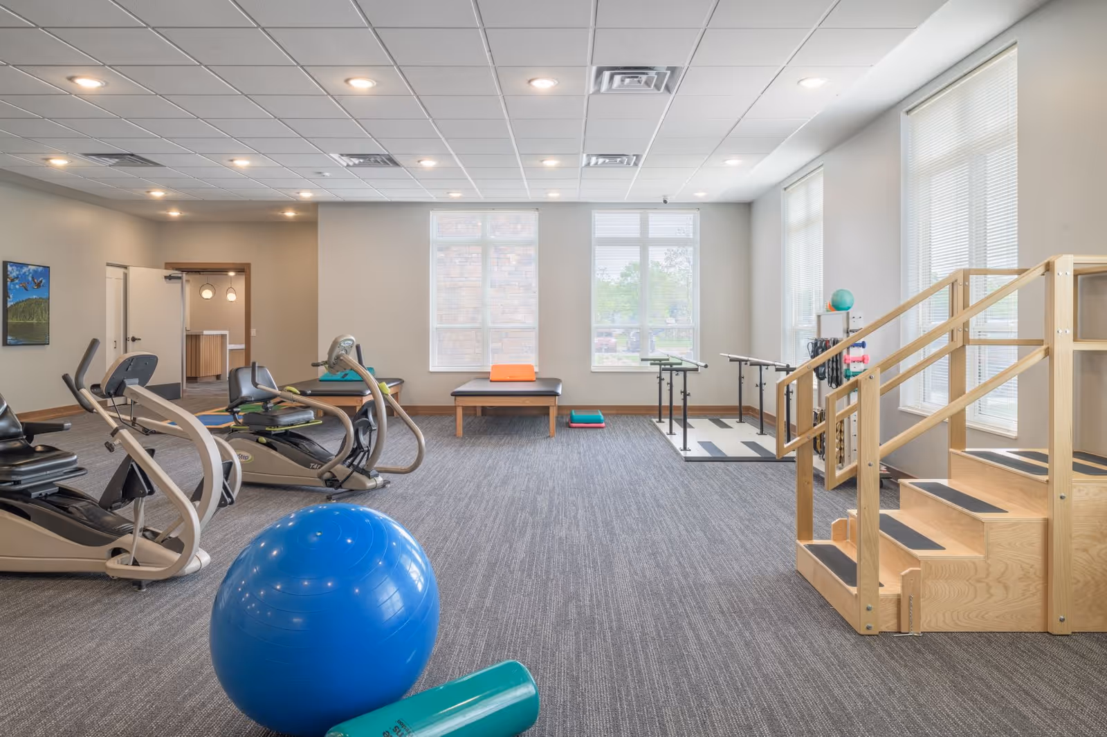 A spacious senior living facility exercise room with large windows allowing natural light. The room contains exercise equipment including stationary bikes, a large blue exercise ball, a green foam roller, a wooden staircase for physical therapy, parallel bars, and a padded bench with exercise mats nearby. The walls are light-colored and the floor is carpeted.