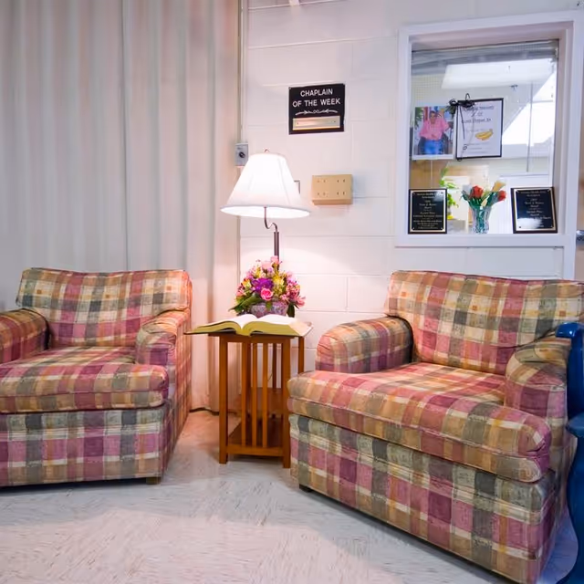 Two plaid armchairs flank a small wooden side table with a lamp, flowers, and an open book in a facility sitting area.