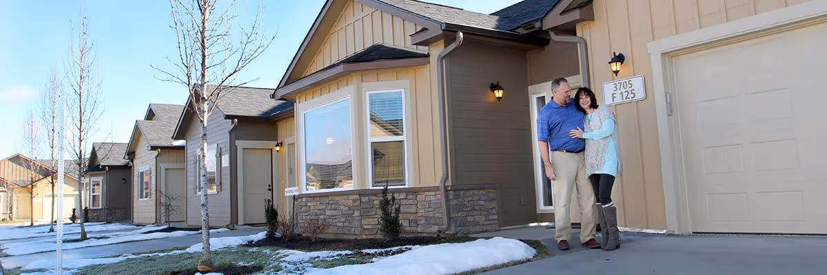 A couple stands outside a beige and brown single-story residential building with a garage. There is snow on the ground and leafless trees along the sidewalk. The man and woman are smiling and embracing near the garage door.