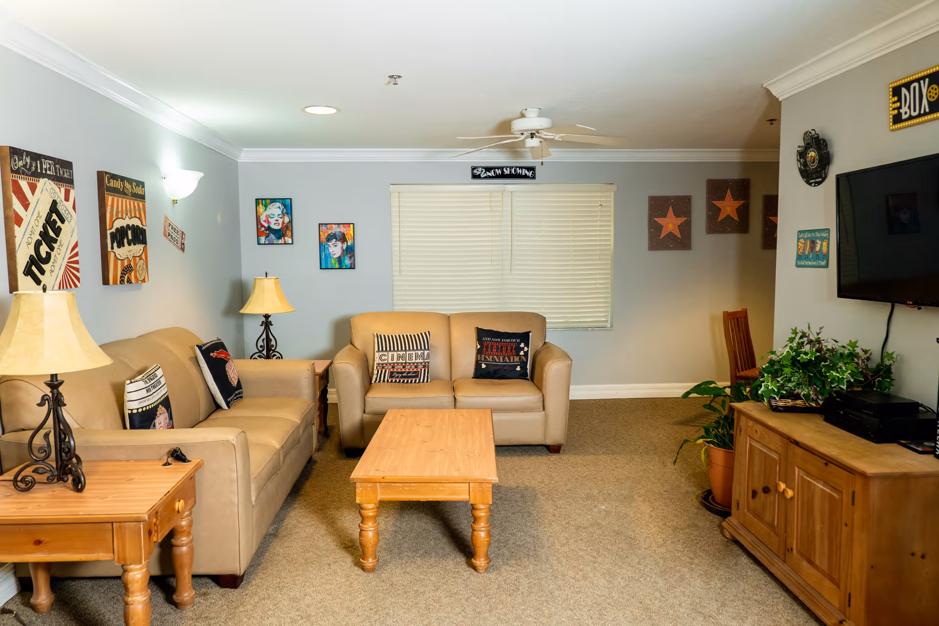 A cozy living room with beige sofas, a wooden coffee table, and side tables with lamps. The walls are decorated with colorful artwork and movie-themed signs. A flat-screen TV is mounted on the wall above a wooden cabinet with plants on top. The room has carpeted flooring and a ceiling fan.