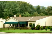 Single-story residential building with a covered porch and green awning, surrounded by a small fence and landscaped bushes, with a backdrop of trees and hills.