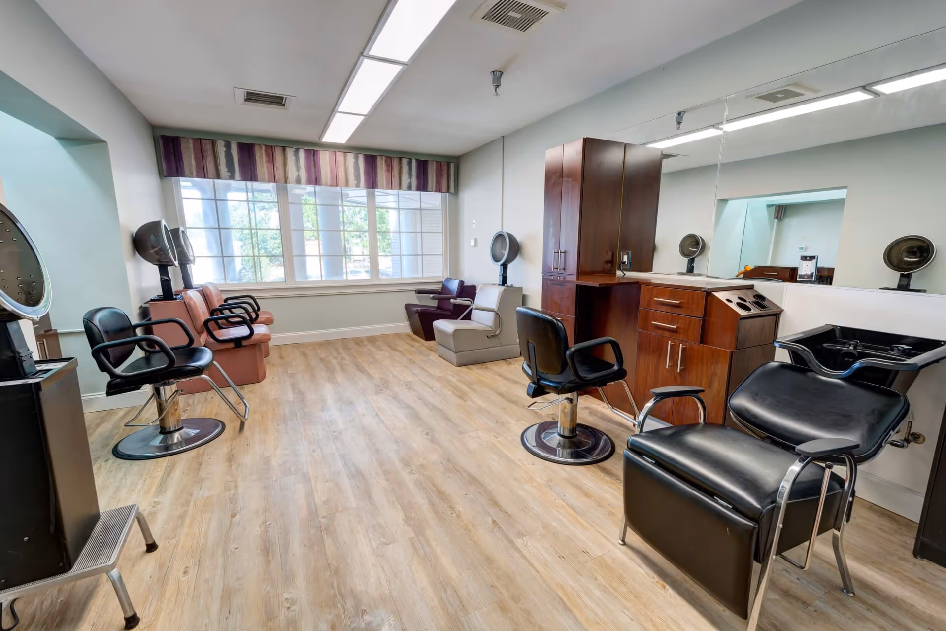 Interior view of a salon area in Ridgeland Place featuring multiple salon chairs, hair dryers, a washing station, wooden cabinets, and a large window with striped valance allowing natural light to fill the room.