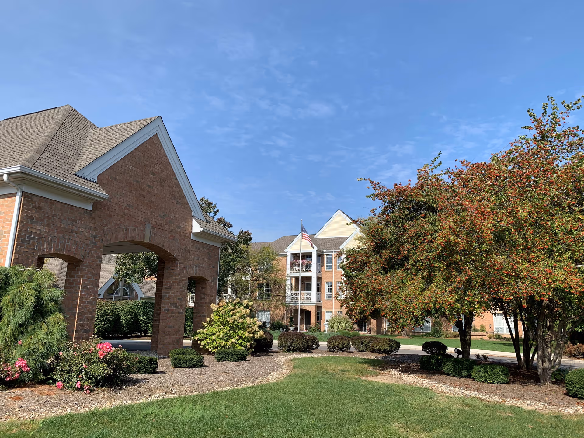Exterior view of The Parkvue Community showing a brick building with a covered entrance, landscaped garden with bushes and flowering plants, and a tree with red berries under a clear blue sky.