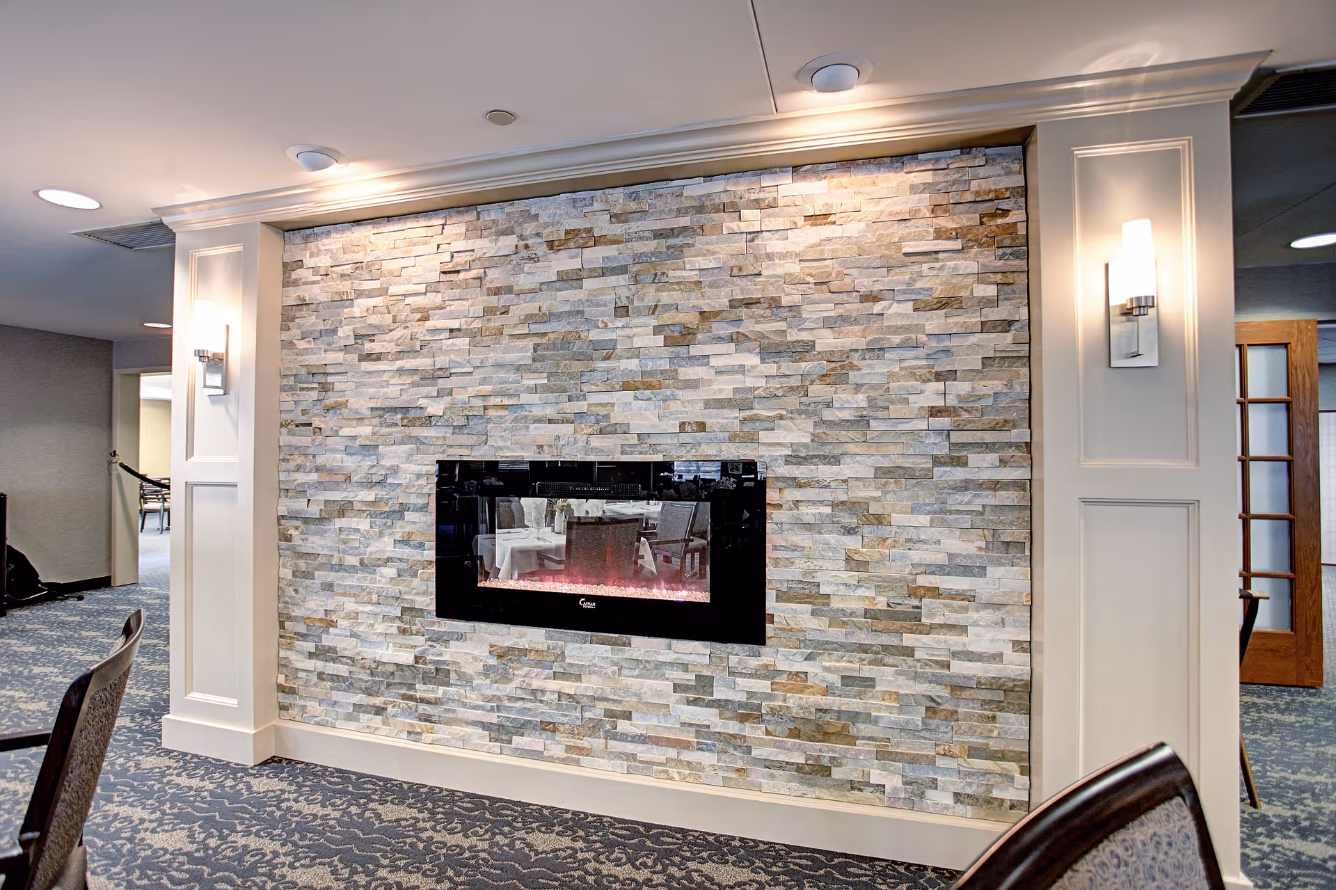 Interior view of a room featuring a modern electric fireplace set into a decorative stone wall. The room has patterned carpet flooring, white paneled walls with sconces on either side of the fireplace, and visible dining chairs and tables reflected in the fireplace glass.
