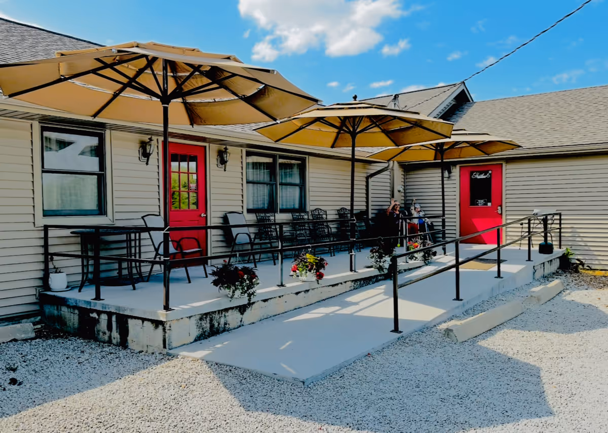 Outdoor patio area at a residential care center with beige umbrellas, black metal chairs, and tables. The patio has a concrete ramp with black railings and flower pots hanging along the railing. The building exterior is beige with two red doors and windows. Two people are seated on the patio under the umbrellas.