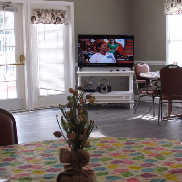 A common area in an assisted living facility with a television on a white stand showing a talk show. There are chairs and tables around, one table covered with a colorful tablecloth and a decorative centerpiece. Large windows and a glass door let in natural light.