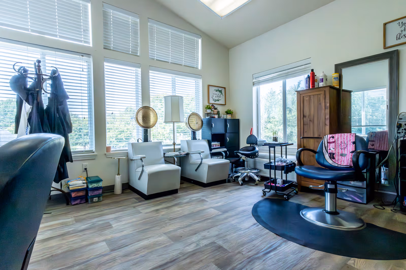 A bright room with large windows covered by white blinds, featuring two white salon chairs with hair dryers above them, a black salon chair with a pink towel draped over it, a tall mirror, a wooden cabinet, and a small rolling cart with hair styling tools. The floor is wood-patterned, and there are plants and framed wall art adding decoration.