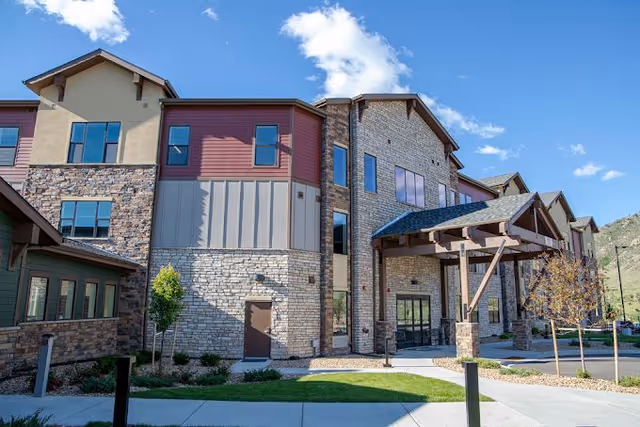 Exterior view of a multi-story assisted living facility building with stone and wood siding, large windows, and a covered entrance under a blue sky with some clouds.
